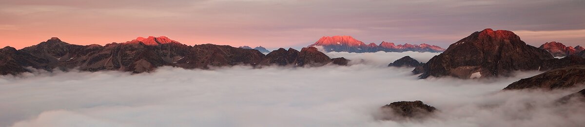 Veduta panoramica delle Alpi Marittime al tramonto, con le cime illuminate da una luce rosata e un mare di nuvole che copre le valli sottostanti; sullo sfondo si distingue il massiccio dell’Argentera. Fine descrizione dell’immagine. Archivio APAM.