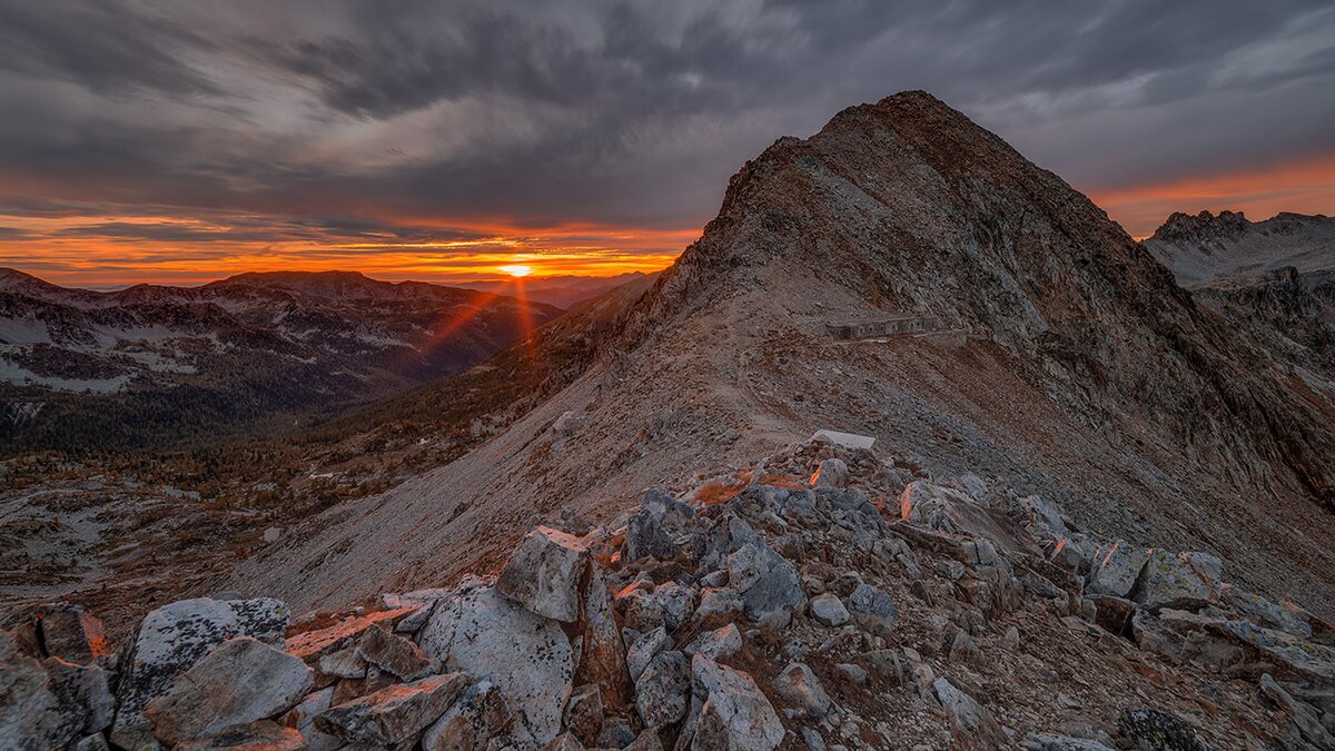 Veduta del colle transfrontaliero di Fremamorta, con il sole che illumina di luce aranciata le rocce e il sentiero di cresta. Foto di M. Lucotti. Fine descrizione dell’immagine.