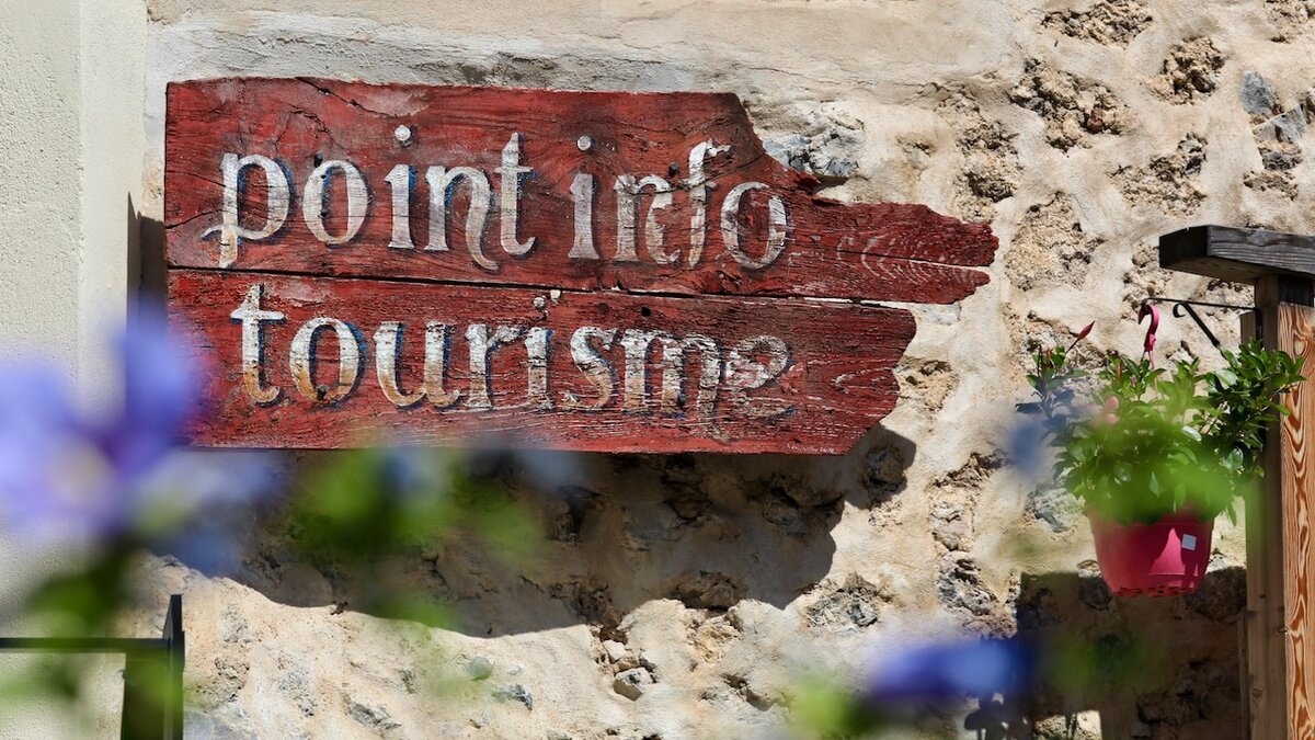 Insegna turistica in legno rosso con la scritta “point info tourisme”, montata su un muro in pietra, con fiori sfocati in primo piano. Fine descrizione dell’immagine.