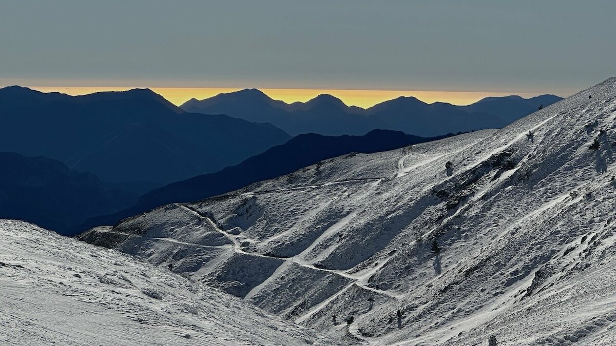 Veduta invernale delle Alpi Marittime con pendii innevati attraversati da un sentiero che serpeggia tra le rocce, sullo sfondo le sagome scure delle montagne al tramonto con una sottile fascia di luce dorata all’orizzonte. Archivio APAM. Fine descrizione dell’immagine.